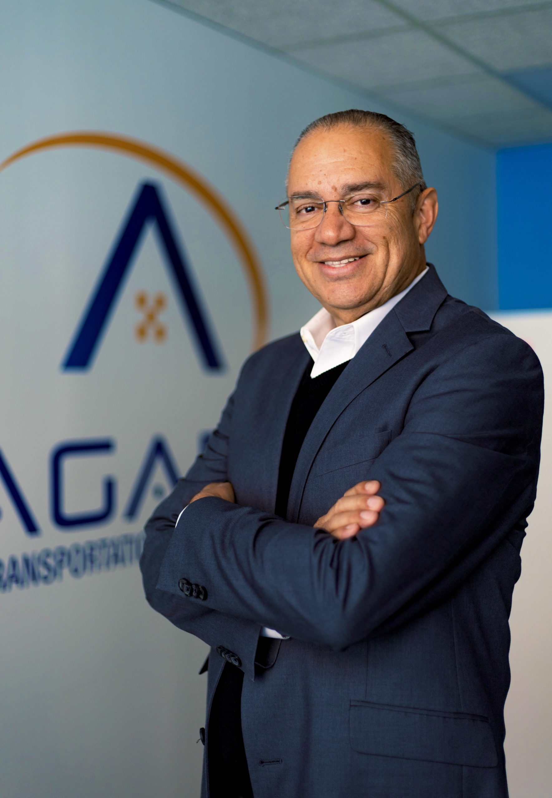 Image of Mario Sena, immigrant founder, president, and CEO of Agape Transportation, standing confidently in front of the Agape Transportation logo. He wears a blue suit jacket over a white shirt and black sweater. This visual embodies our commitment to excellence in non-emergency medical transportation, serving New York City. Mario Sena's leadership mirrors our dedication to providing exceptional medical transportation services