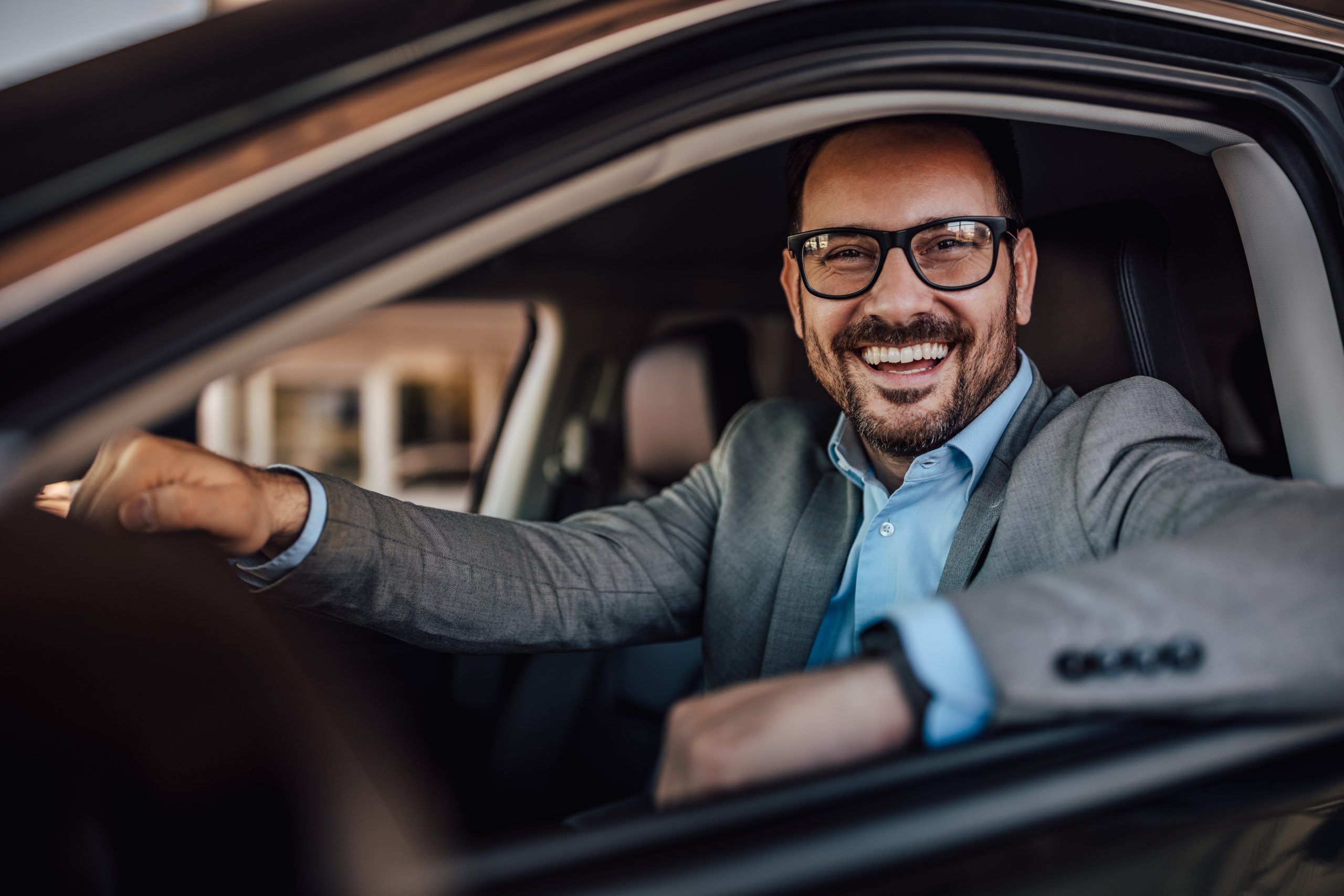 Man with glasses on, dressed in a suit, posing while sitting in his new car.